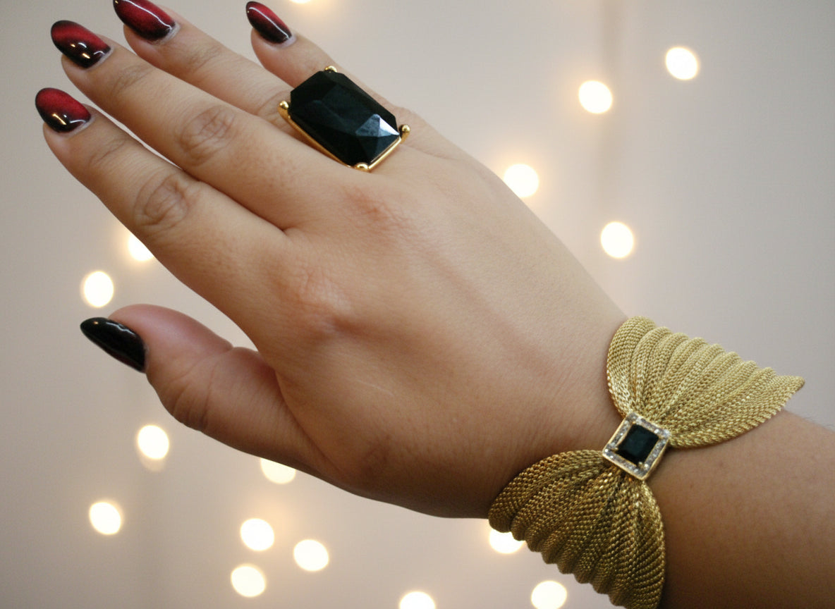 Hand wearing a gold ring and bracelet with black gemstones on a plain background